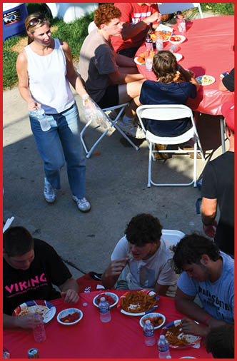 Several people are gathered around red-covered tables outdoors, eating food and interacting, while one adult holds the hand of a child walking nearby. Description generated by AI