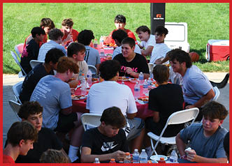 A group of people sitting around a table outdoors on a sunny day, eating and drinking while engaged in conversation. Description generated by AI
