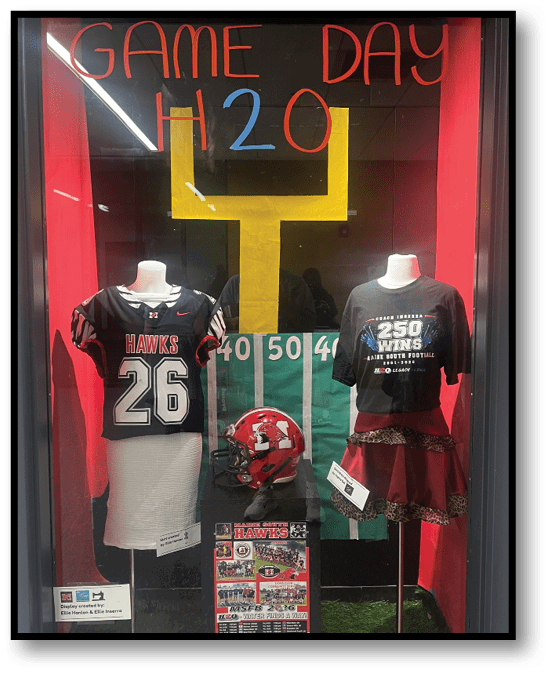 A sports-themed display features a large yellow goalpost with the words “GAME DAY" and "H2O" written above it, flanked by two mannequins wearing sports jerseys. Description generated by AI