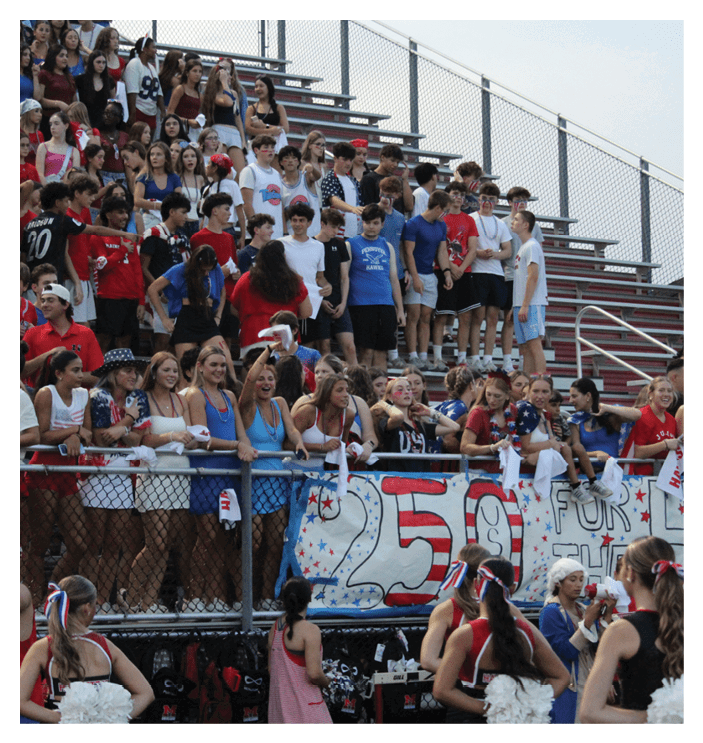 A large group of people wearing casual and patriotic clothing are gathered in bleachers at an outdoor event, some smiling and cheering while holding a decorated banner. Description generated by AI