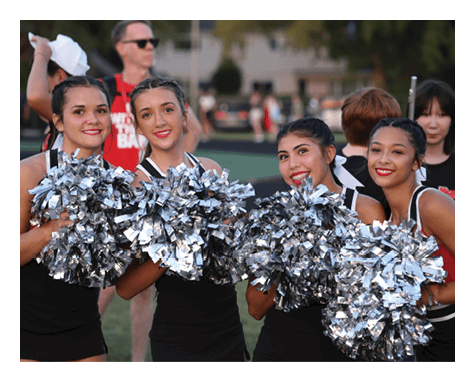 Three smiling adults in cheerleading uniforms hold large silver pom-poms outdoors. Description generated by AI