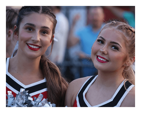 Two adults with braided hair and bright lipstick smile while wearing cheerleading uniforms in an outdoor setting. Description generated by AI