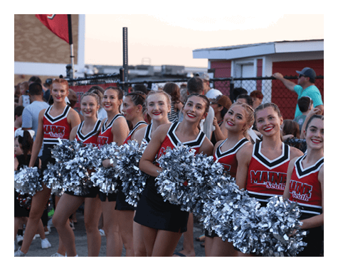A group of smiling adults dressed in red and black cheerleading uniforms hold silver pom-poms while standing outdoors near a fence with other people in the background. Description generated by AI
