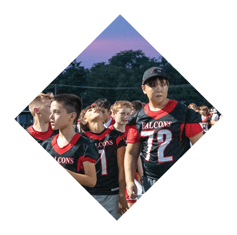 Children wearing Falcons football jerseys are gathered on a field during dusk, some looking around while others appear engaged in conversation. Description generated by AI