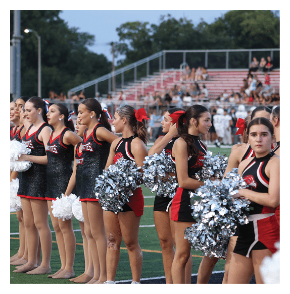 A line of people dressed in matching black and red cheerleading uniforms holding white pom-poms stand on a sports field. Description generated by AI
