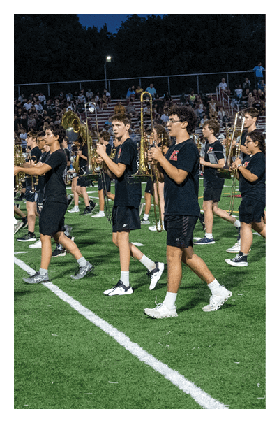 People wearing black shirts and shorts are marching on a football field at night while playing trombones with a crowd seated in bleachers in the background. Description generated by AI
