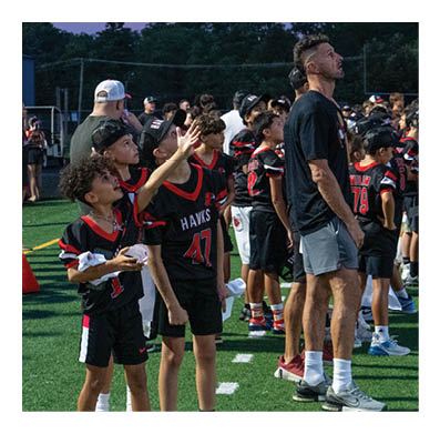 A group of people, including children in sports uniforms and adults in casual clothing, stand in a line on a football field near the sideline during dusk. Description generated by AI
