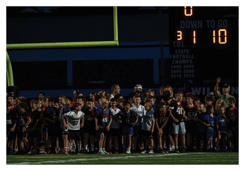 A large group of children wearing sports jerseys are gathered together on a field under a goalpost at night. Description generated by AI