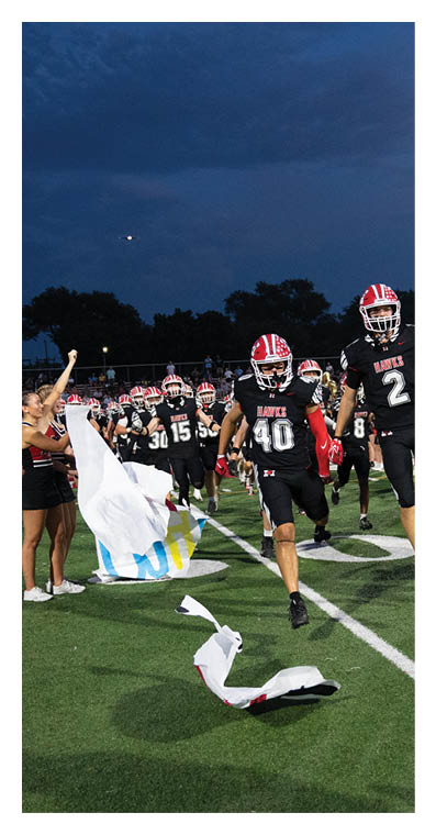 People in football uniforms are running onto a field at dusk while cheerleaders wave a torn banner. Description generated by AI