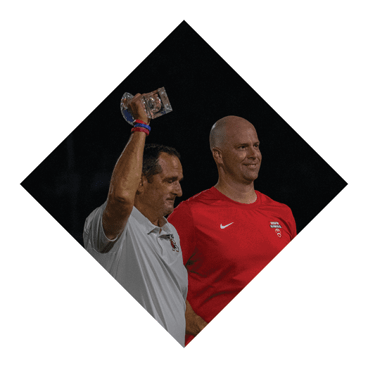 Two adults stand side by side at night, one wearing a red sports shirt and the other raising a hand holding a small object. Description generated by AI
