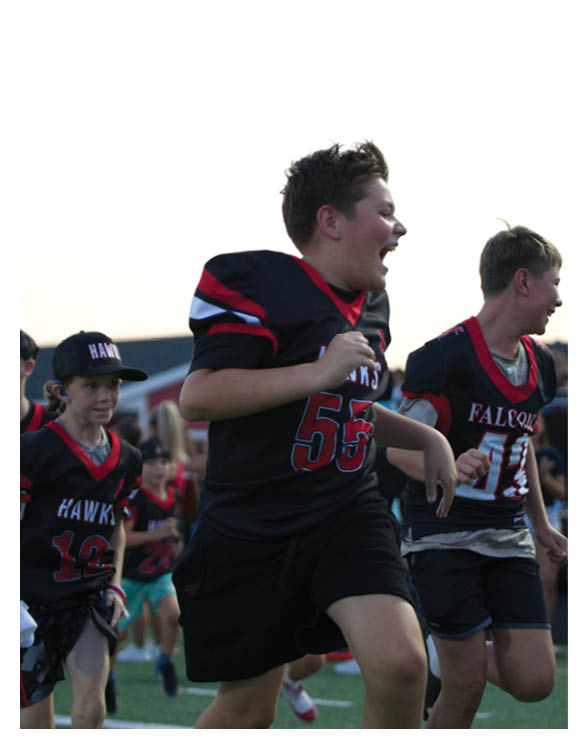 Children wearing sports jerseys are running and appear to be excited or shouting during an outdoor event. Description generated by AI