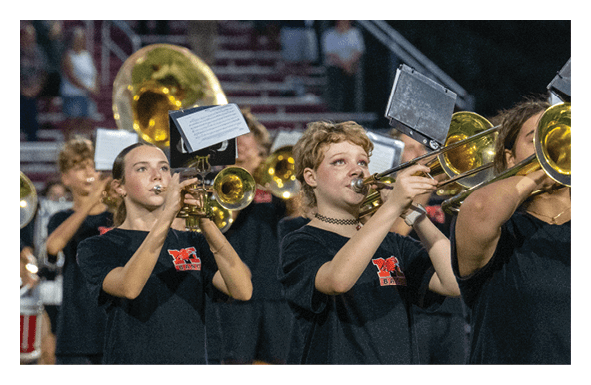 People wearing black shirts with red logos are playing brass instruments in a band setting, with music sheets attached to their instruments. Description generated by AI