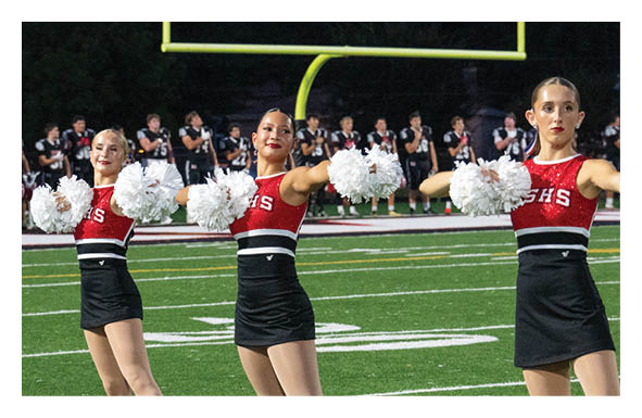 Two adults in red and black cheerleading uniforms hold white pom-poms while performing on a football field with players standing in the background. Description generated by AI