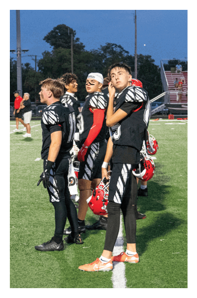 Four people wearing black and white football uniforms stand on a field with helmets in hand during what appears to be a game or practice at dusk. Description generated by AI