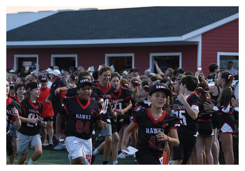 Children wearing sports jerseys run on a field while a group of cheerleaders and other children stand in the background near a red building. Description generated by AI