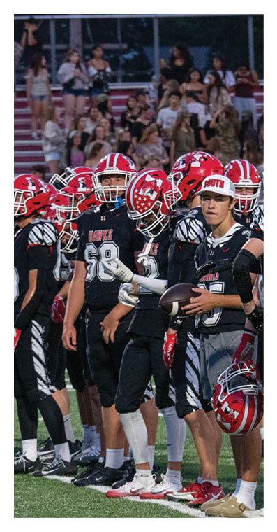 A group of people wearing black football uniforms and red helmets stand together on a field with a crowd in the background. Description generated by AI