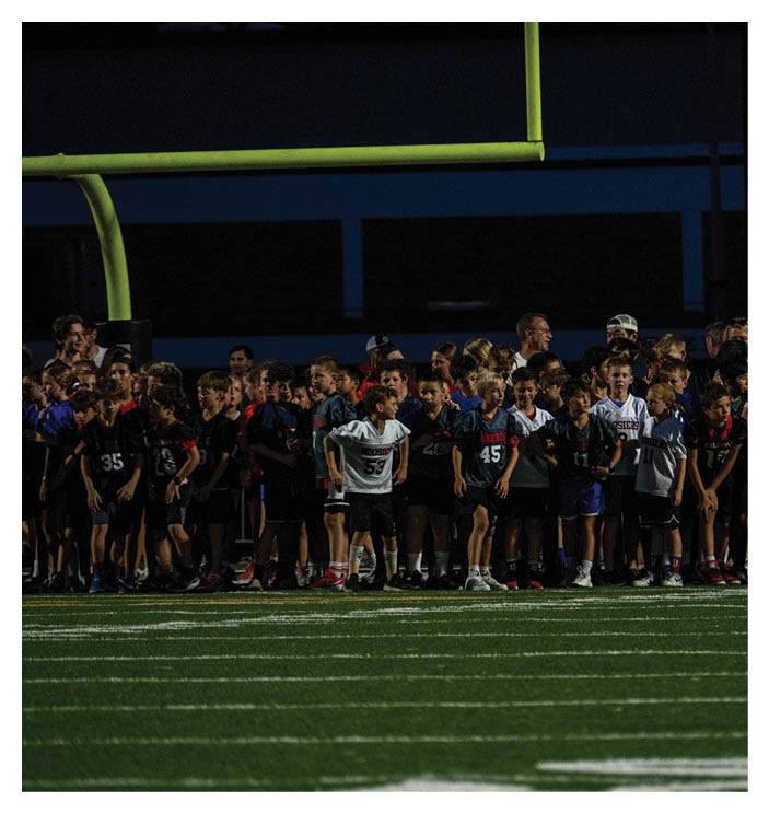 A large group of children wearing sports jerseys stand together on a football field under stadium lights near a goalpost at night. Description generated by AI