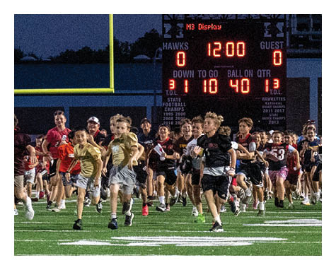 A large group of people wearing sports and casual clothing are running energetically across a football field at dusk with a scoreboard and an American flag visible in the background. Description generated by AI