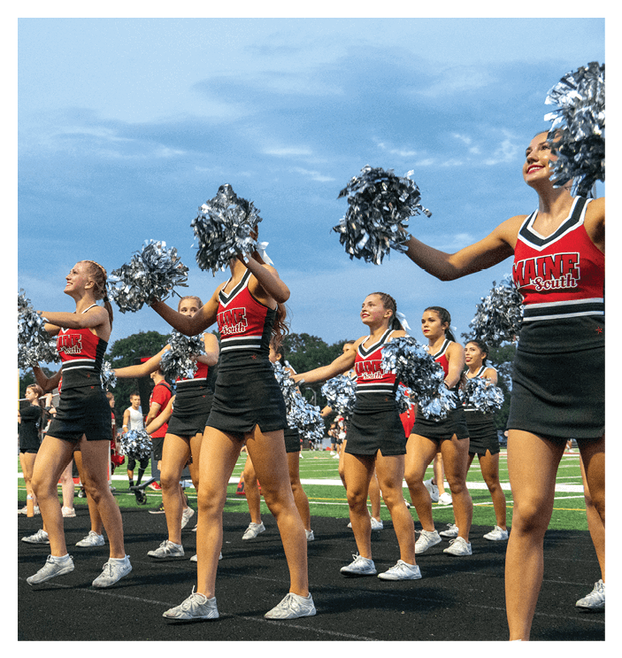 A group of adults in matching cheerleading uniforms with pom-poms perform a routine on a sports field under a cloudy sky. Description generated by AI