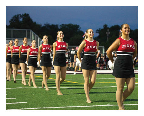 A group of adults in matching red and black cheerleading uniforms are smiling and walking in formation on a sports field during dusk. Description generated by AI
