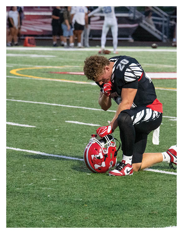 A person in a black and white sports uniform kneels on a football field with a red helmet placed on the ground in front of them while holding their face. Description generated by AI