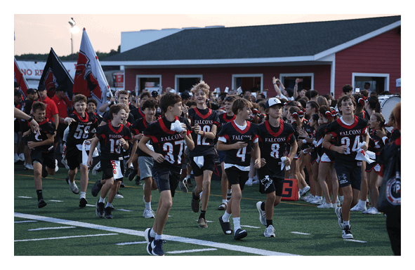 A group of young athletes in black and red football uniforms are running on a field with cheerleaders and a crowd cheering in the background near a red building. Description generated by AI