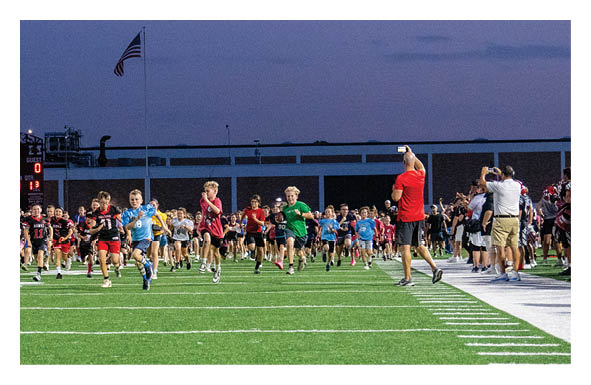 Children wearing colorful athletic clothing are running on a football field at dusk while adults stand on the sidelines watching. Description generated by AI