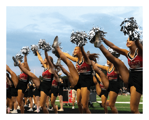 A group of people in matching cheerleading uniforms are performing a high kick routine on a sports field while holding pom-poms. Description generated by AI