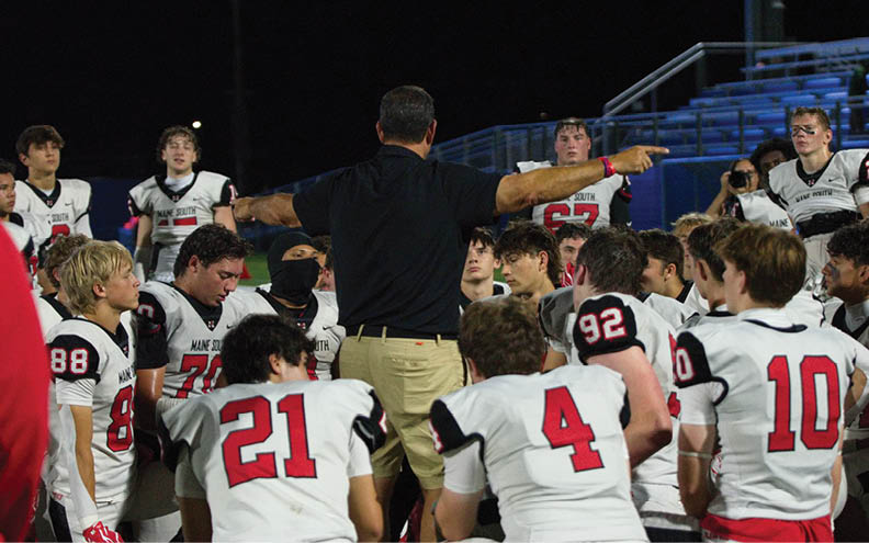 A group of people in football uniforms are gathered around a coach who is giving instructions on a field at night. Description generated by AI