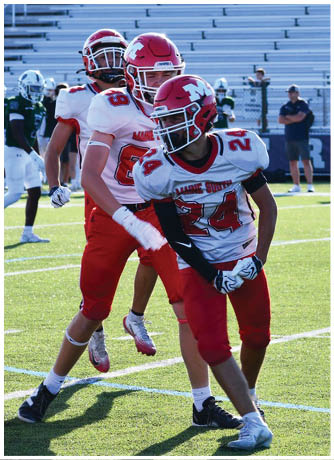 Two people in red and white football uniforms are on a green field with one person preparing to catch or throw a football while the other stands nearby. Description generated by AI