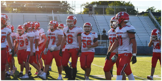 Several people wearing red and white football uniforms and helmets are standing together on a sunlit field with empty bleachers in the background. Description generated by AI