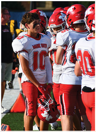 Three people wearing red football helmets and white jerseys with red lettering are gathered closely, with one person having black face paint under their eyes. Description generated by AI