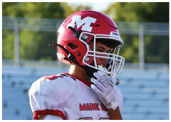 A person wearing a red football helmet with a white “M" and a white football jersey is adjusting their helmet on a field with blurred bleachers and trees in the background. Description generated by AI