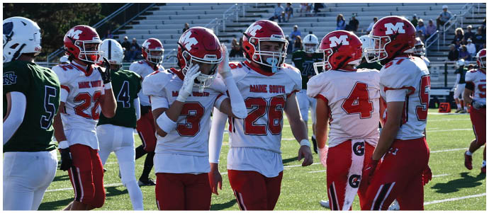 People in football uniforms from two teams are standing on a field with bleachers and spectators in the background. Description generated by AI
