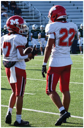 Two people wearing red and white football uniforms stand on a green field with one holding a football. Description generated by AI
