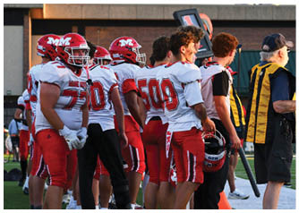 A group of people wearing red and white football uniforms stand together on a field near a building during daylight. Description generated by AI