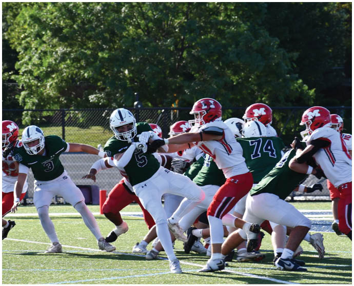 People in football uniforms are actively engaged in a game on a grassy field with trees and a fence in the background. Description generated by AI