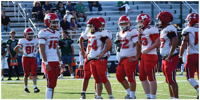 Several people wearing red and white football uniforms stand on a green field with bleachers in the background. Description generated by AI