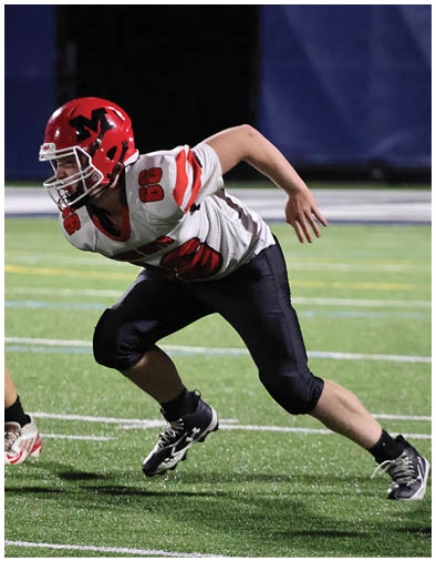 A person wearing a red helmet, white jersey with red stripes, and black pants is crouched and running on a football field. Description generated by AI