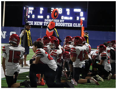 A group of people wearing red and white football uniforms are kneeling on a field near two officials holding orange down markers during a nighttime game. Description generated by AI