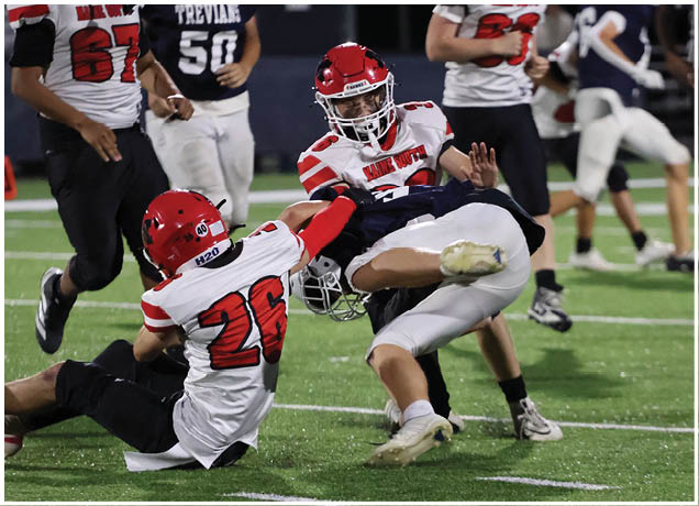 Two people in football uniforms are engaged in a tackle on a grassy field during a game with other players and a referee visible in the background. Description generated by AI