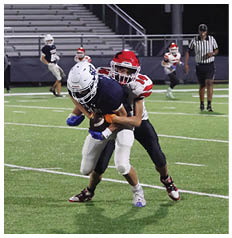 A person in a red helmet and white and red sports uniform is tackling another person in a dark uniform on a football field at night with a referee and another player in the background. Description generated by AI