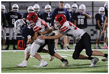 Two football players in white and red uniforms tackle a player in a dark uniform on a field with spectators and coaches in the background. Description generated by AI