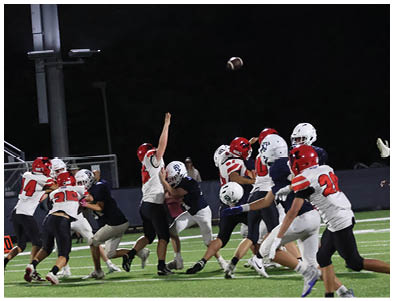 A group of people wearing football helmets and uniforms are actively playing on a field at night with a football in mid-air. Description generated by AI