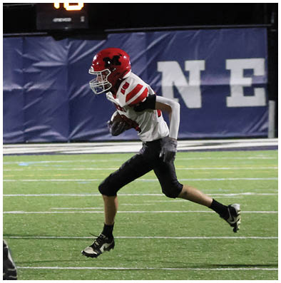 A person wearing a red helmet and black pants is running on a football field while holding a football, with two other people in sports uniforms nearby. Description generated by AI