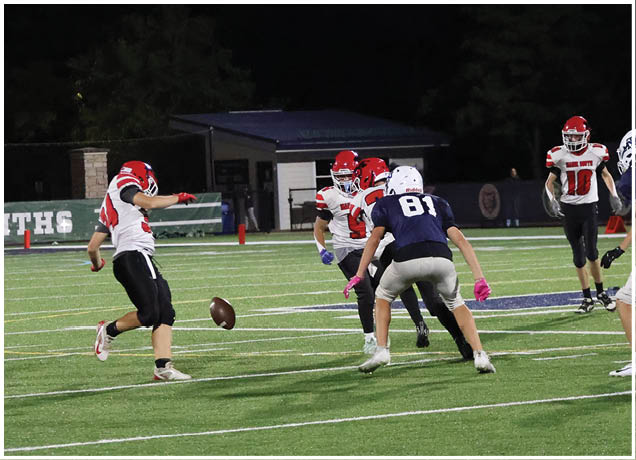 Players in red and white and blue and white uniforms are actively engaged in a nighttime football game on a green field with one player kicking the ball. Description generated by AI