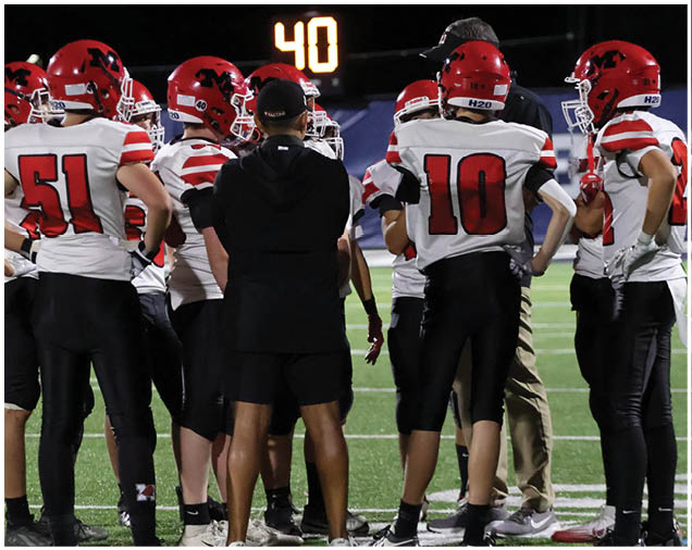 A group of people wearing red helmets and white sports jerseys with black pants are gathered on a football field under stadium lights. Description generated by AI