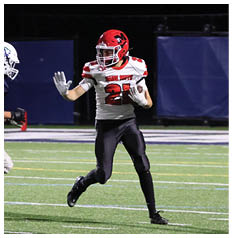 A person wearing a red helmet and white and red football uniform is running on a football field while holding a football and raising one hand. Description generated by AI
