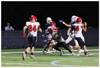 Several people wearing football uniforms are actively engaged in a play on a well-lit field at night. Description generated by AI