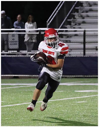 A person wearing a red helmet and white and red football uniform is running on a green football field while holding a football. Description generated by AI